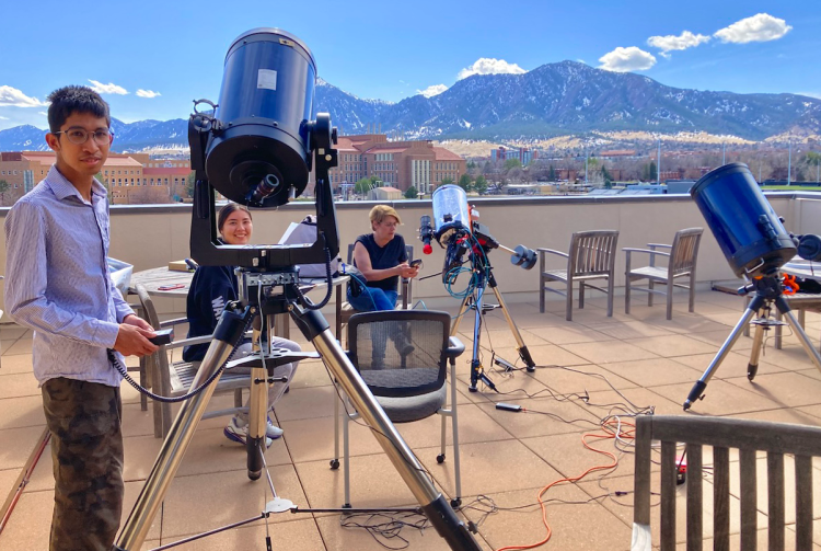 Three people gather on a roof to assemble three telescopes. Mountains in the background